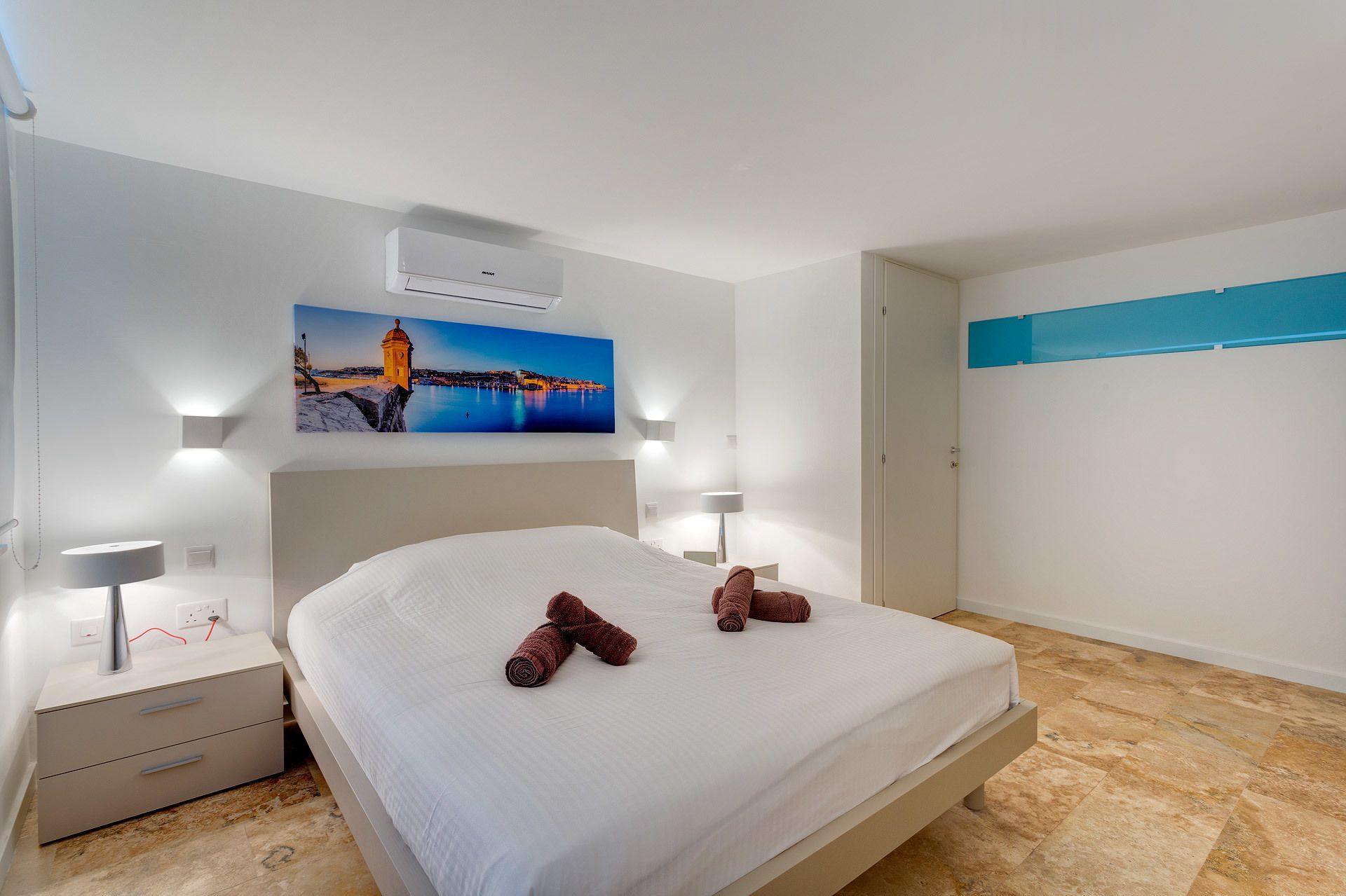 A hotel room with a bed, a nightstand, a lamp, a wall-mounted air conditioner, and a painting of a coastal scene.