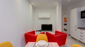 A modern dining area with a table set for two, red and yellow chairs, a flat-screen TV, and a white cabinet above the TV. - Cozy Apartment in an Amazing Location (Sliema)