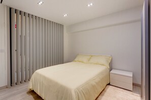 A bedroom with a bed, a nightstand, a white dresser, and a sliding door with vertical blinds.