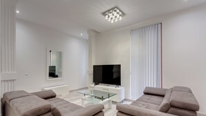 Ein modernes Wohnzimmer mit Fernseher, zwei beigefarbenen Sofas, einem Glastisch und einem großen Fenster mit Jalousien.
