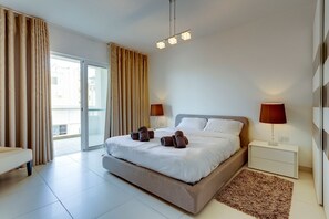 A modern hotel room with a large bed, two bedside lamps, a small bedside table, and a view of the outside through a sliding glass door. - Contemporary, Luxury Apartment With Valletta and Harbour Views (Sliema)