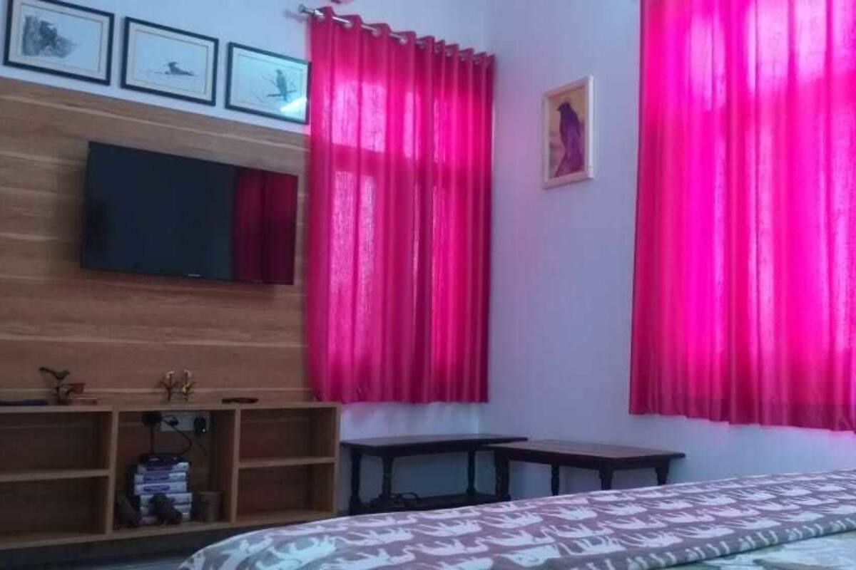 Une chambre avec un lit, un téléviseur fixé au mur, une étagère en bois avec quelques objets et des rideaux roses.