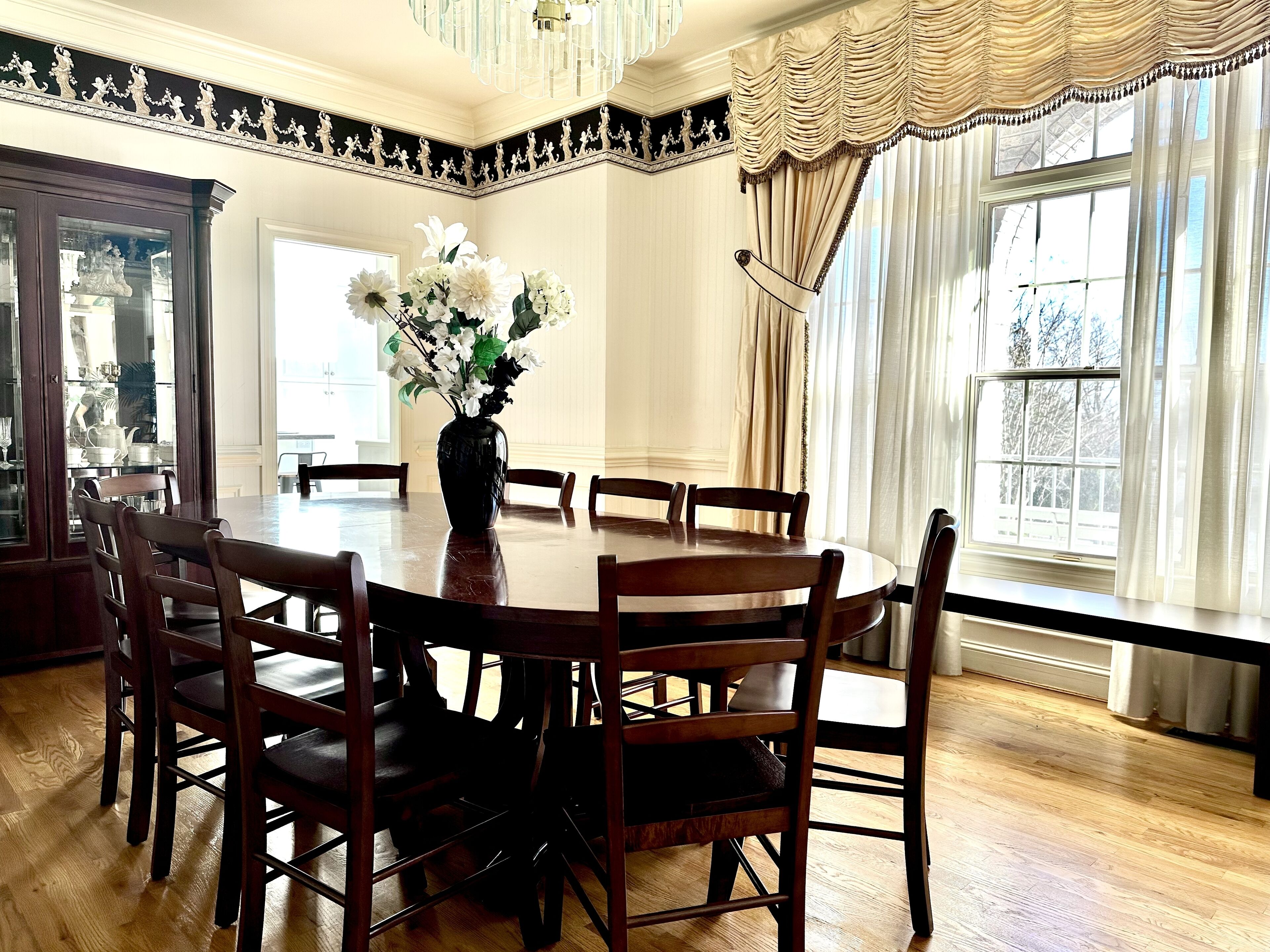 A dining room with a round wooden table, six chairs, and a vase with white flowers.
