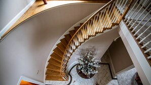 Una imponente escalera de caracol con escalones de madera y barandillas blancas, que conduce a un rellano con suelo de mármol y un arreglo floral.