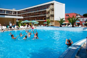 Zona de piscina con sillones de descanso y sombrillas, un edificio de varios pisos y balcones, y un cielo azul despejado.