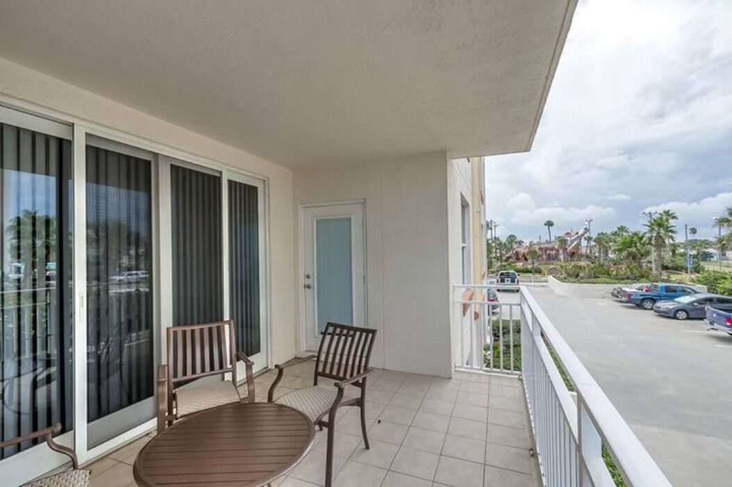 A balcony with a table and chairs, a sliding glass door, and a view of a parking area with cars and palm trees.