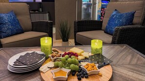 Un espace extérieur accueillant avec des meubles en osier et des coussins, une table avec un plateau de fromages et des amuse-gueules, et une vue sur une enseigne lumineuse d’une salle d’arcade en arrière-plan.