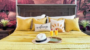 Un lit avec une couvre-lit jaune, plusieurs oreillers, un oreiller décoratif avec l’inscription « Bienvenue », deux verres de jus d’orange et un chapeau blanc, le tout posé sur un plateau.