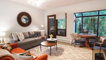 A living room with a sofa, a coffee table, a dining table with chairs, a large window, and a decorative mirror.