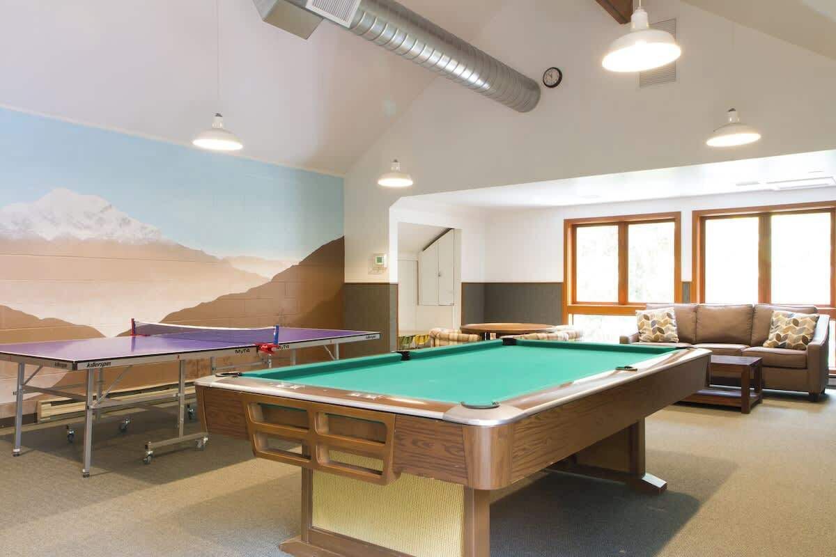 A pool table, ping pong table, and a sofa in a room with a mountain mural and large windows.
