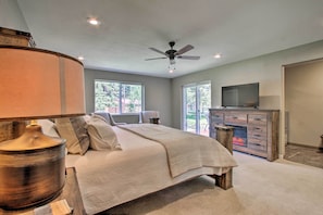 A bedroom with a bed, a ceiling fan, a television, a fireplace, and large windows.