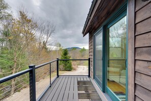 Una terraza de madera con baranda de metal, puerta de cristal y vista a un área boscosa con un sendero de tierra.