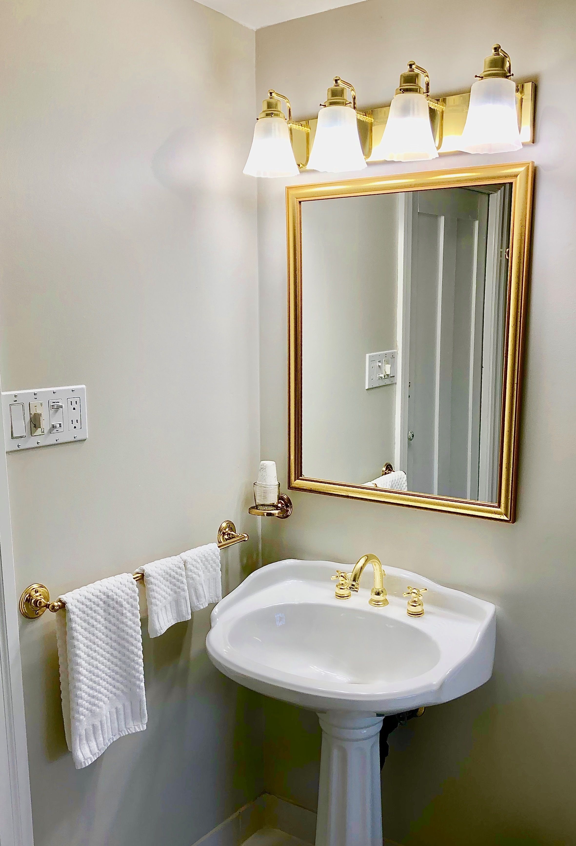 Casa de banho com lavatório branco, acessórios dourados e um espelho grande com moldura dourada.