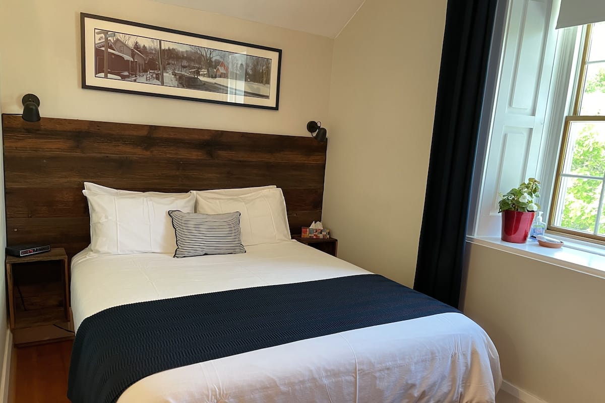 A neatly made bed with white and striped pillows, a dark blue blanket, a wooden headboard, a bedside table with a lamp, a framed picture on the wall, and a window with a plant.
