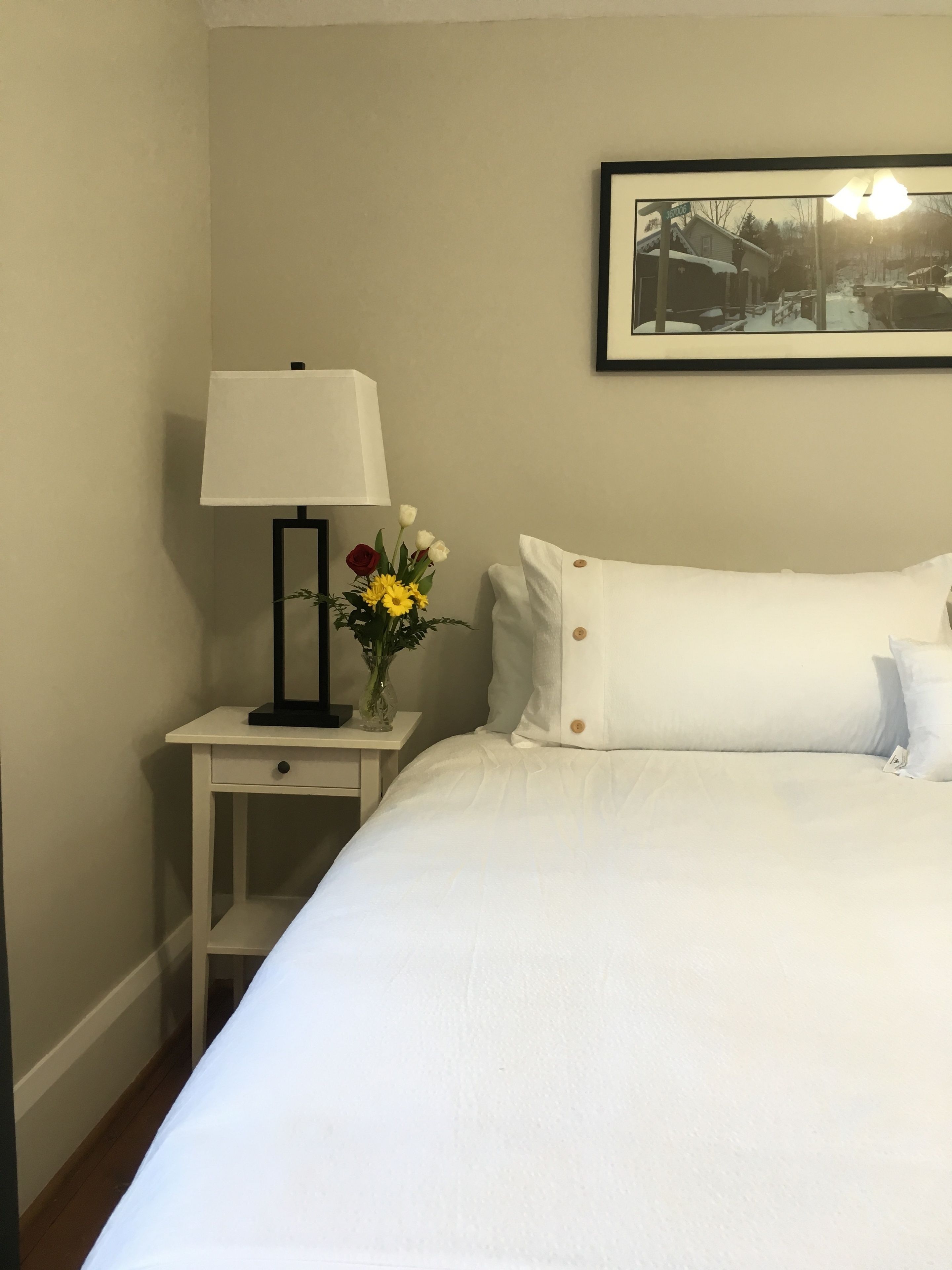 A neatly made bed with white linens, a bedside table with a lamp and a vase of flowers, and a framed picture on the wall.