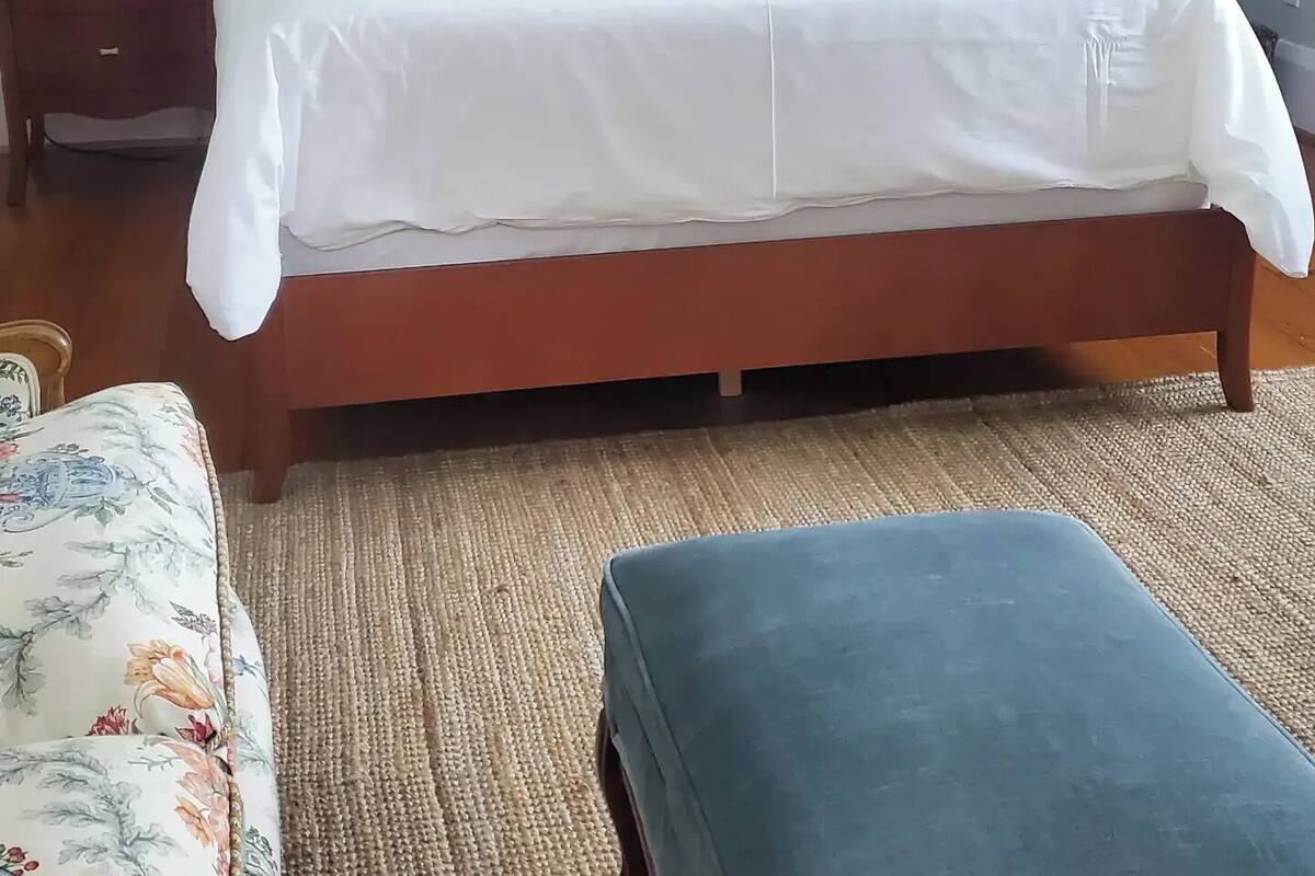 A neatly made bed with white bedding and a single mustard-colored pillow, flanked by two bedside tables with lamps, a framed picture on the wall, and a small bedside table with a tissue box.