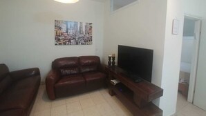 Una sala de estar con un sofá de cuero marrón, un televisor de pantalla plana sobre un soporte de madera, una lámpara de techo y un cuadro de un paisaje urbano en la pared.