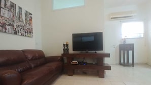 Una sala de estar con un sofá de cuero marrón, un televisor de pantalla plana sobre un soporte de madera, un aire acondicionado montado en la pared y una ventana con persianas.