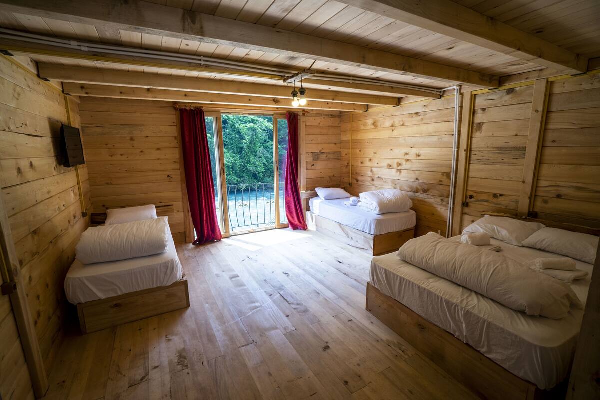 Une cabane en bois avec deux lits simples, une petite table et une fenêtre ornée de rideaux rouges.