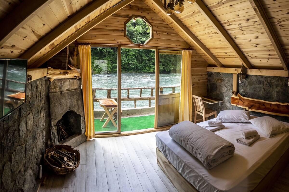Une cabane en bois avec une chambre à coucher située en mezzanine, un mur en pierre, un banc en bois et une vue sur une rivière.