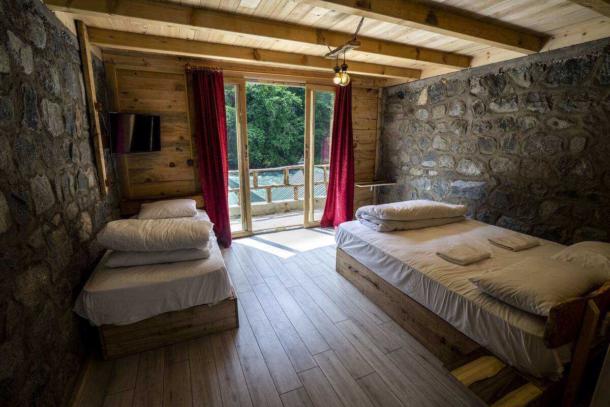 Une cabane avec deux lits en bois, des murs en pierre, un ventilateur de plafond et une vue sur l’extérieur.