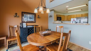 Un comedor con mesa de madera, sillas, una lámpara de araña y una cocina al fondo.