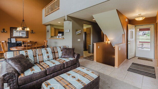 Una sala de estar con sofá, reposapiés, comedor, cocina y una escalera.
