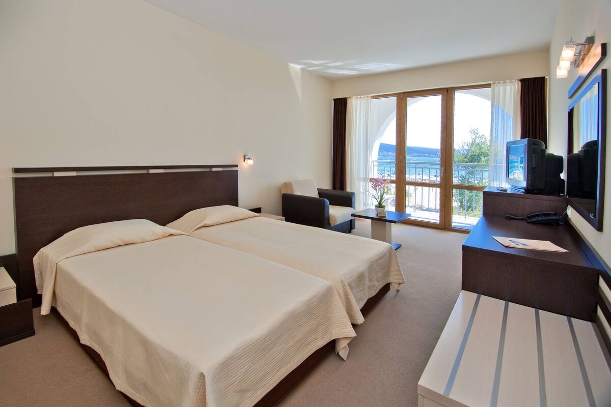 Ein Hotelzimmer mit zwei Betten, einem Schreibtisch mit Computer, einem Fernseher und einem Balkon mit Blick.