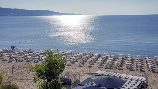 A beach with rows of umbrellas and sun loungers, a fenced area with a small building, and a clear view of the sea. - Hotel Viand (Sunny Beach)