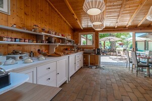 Un interior de madera con mostrador de cocina, estantes con frascos y una zona de comedor con sillas y sombrillas.