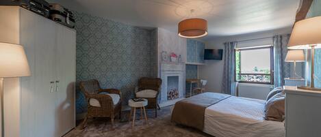 A bedroom with a bed, two wicker chairs, a small table, a wardrobe, a fireplace, and a window with curtains.