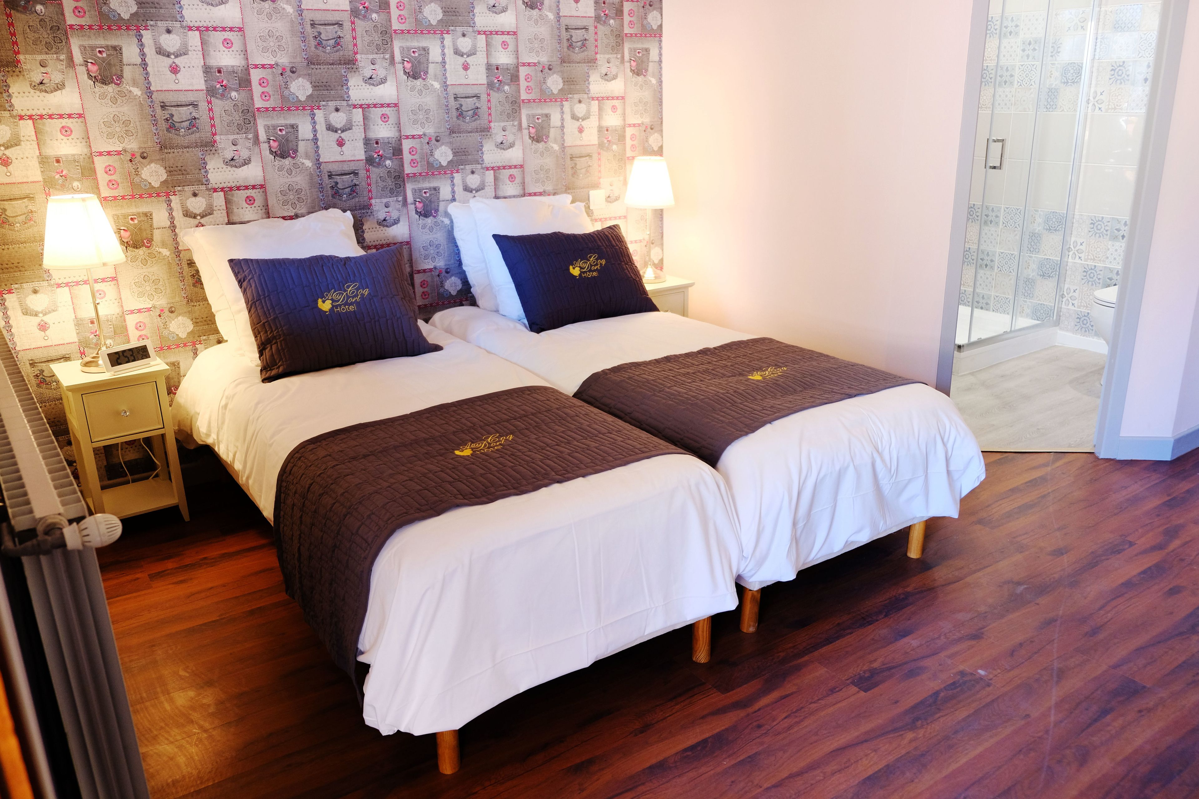 A hotel room with two beds, a patterned wallpaper, a wooden floor, and a glass shower enclosure.