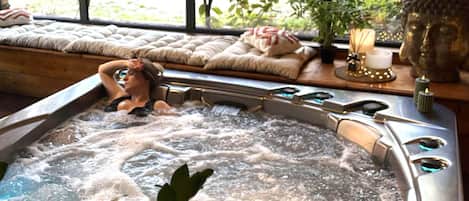 Un jacuzzi con una persona relajándose, una banca acolchada, una estatua de Buda y una vela sobre una mesa.