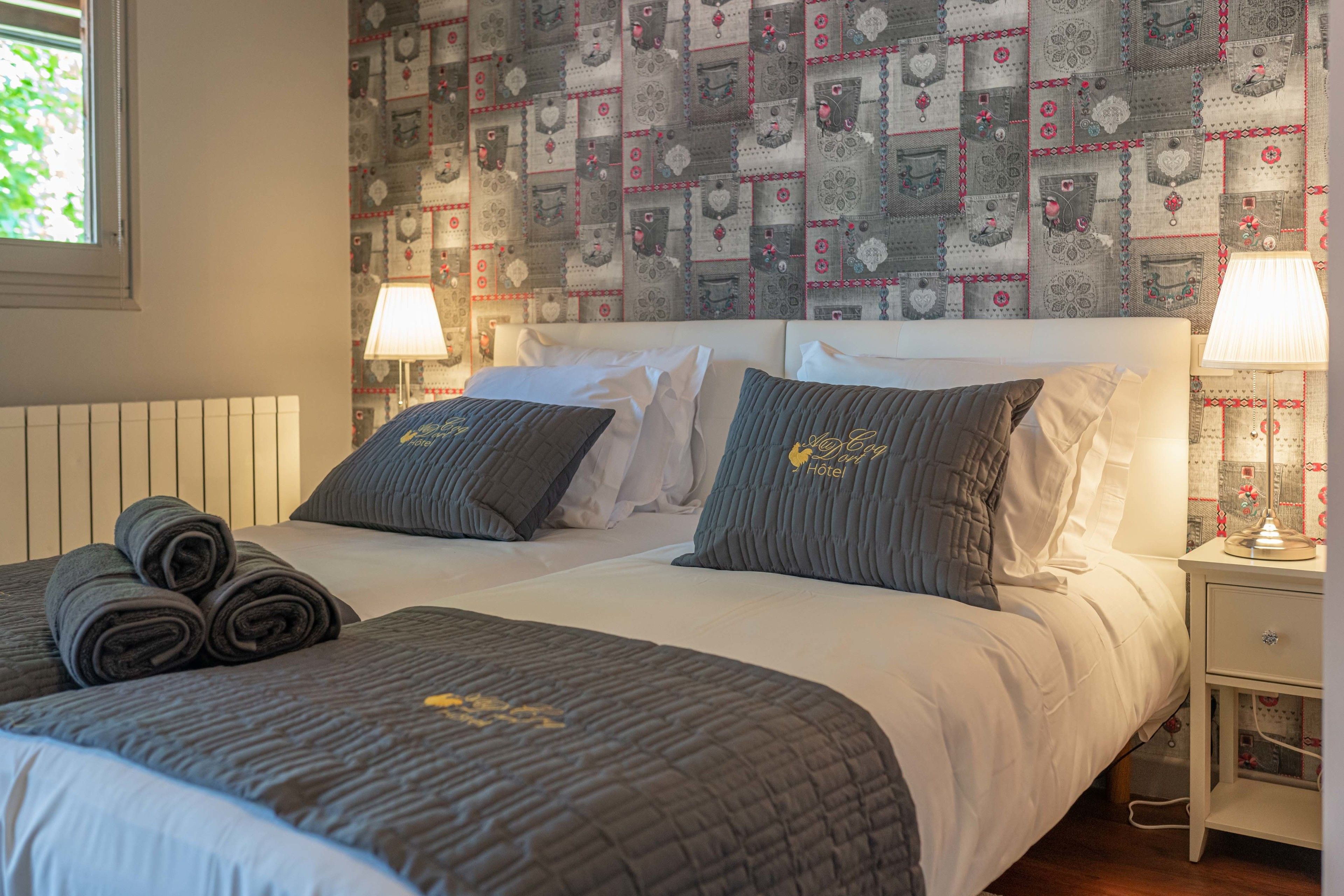 Una cama bien hecha con una colcha gris acolchada, dos almohadas y toallas enrolladas al lado. Hay una mesita de noche con una lámpara y un papel tapiz estampado detrás de la cama.