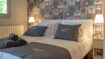 Una cama bien hecha con una colcha gris acolchada, dos almohadas y toallas enrolladas al lado. Hay una mesita de noche con una lámpara y un papel tapiz estampado detrás de la cama.