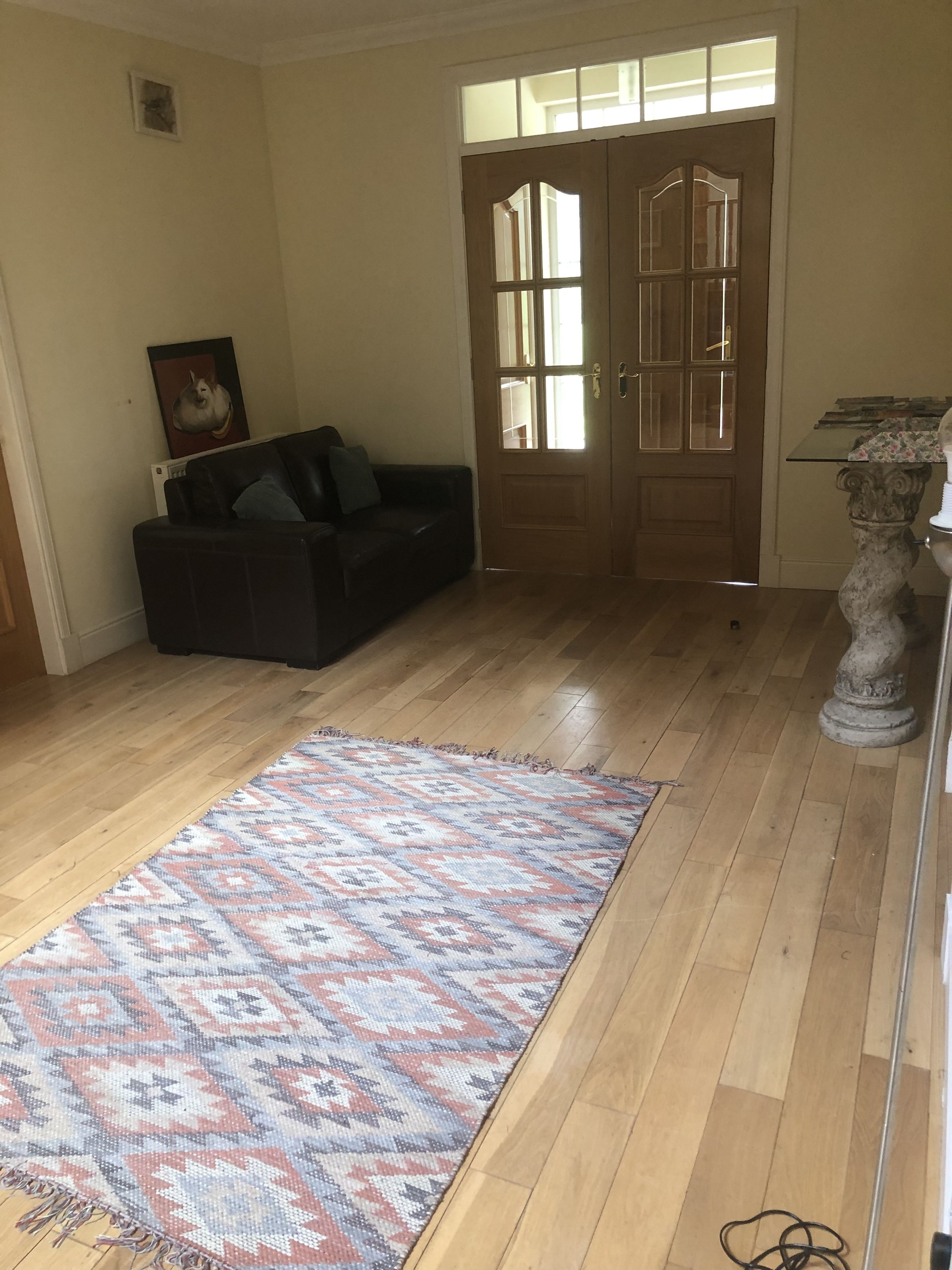 Een kamer met houten vloer, een kamerbreed tapijt met dessin, een zwarte bank, een deur met glaspanelen en een klein tafeltje met decoratieve objecten.