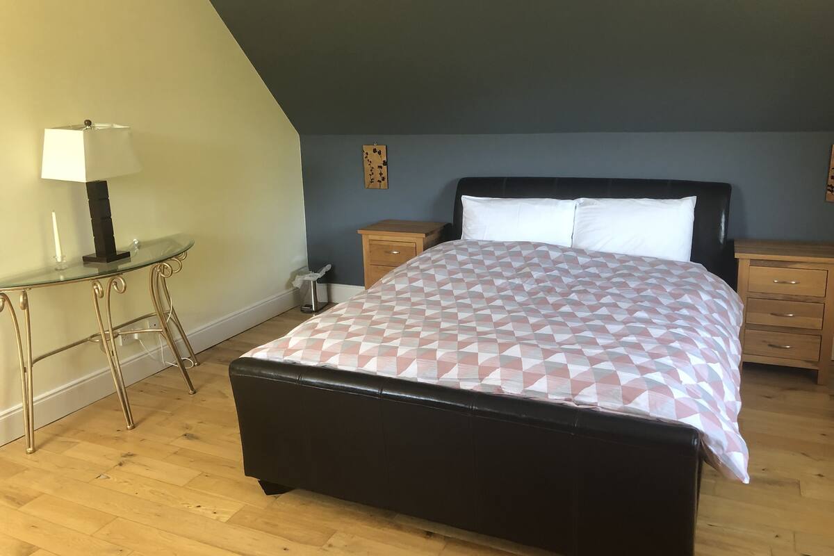 Un dormitorio con cama, mesita de noche, una pequeña mesa con lámpara y una obra de arte en la pared.