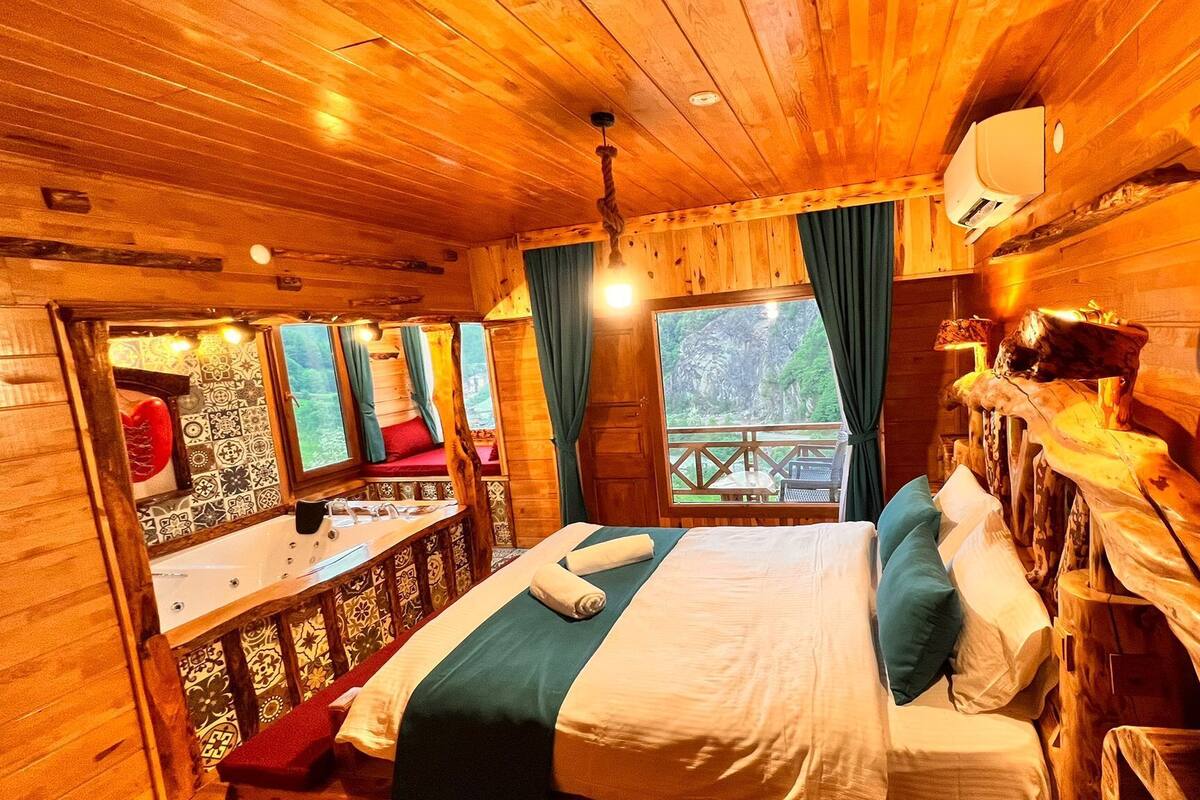 Une chambre dans une cabane en bois, avec un lit, une fenêtre avec des rideaux, une petite table avec une lampe, et une vue sur l’extérieur.