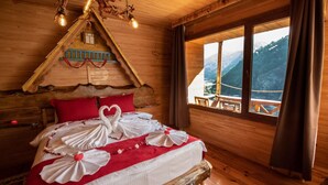 Une chambre confortable dans un chalet en bois, avec un lit décoré de motifs en forme de cœur, un plafond en bois agrémenté de luminaires suspendus et une fenêtre donnant sur une vue imprenable sur les montagnes.