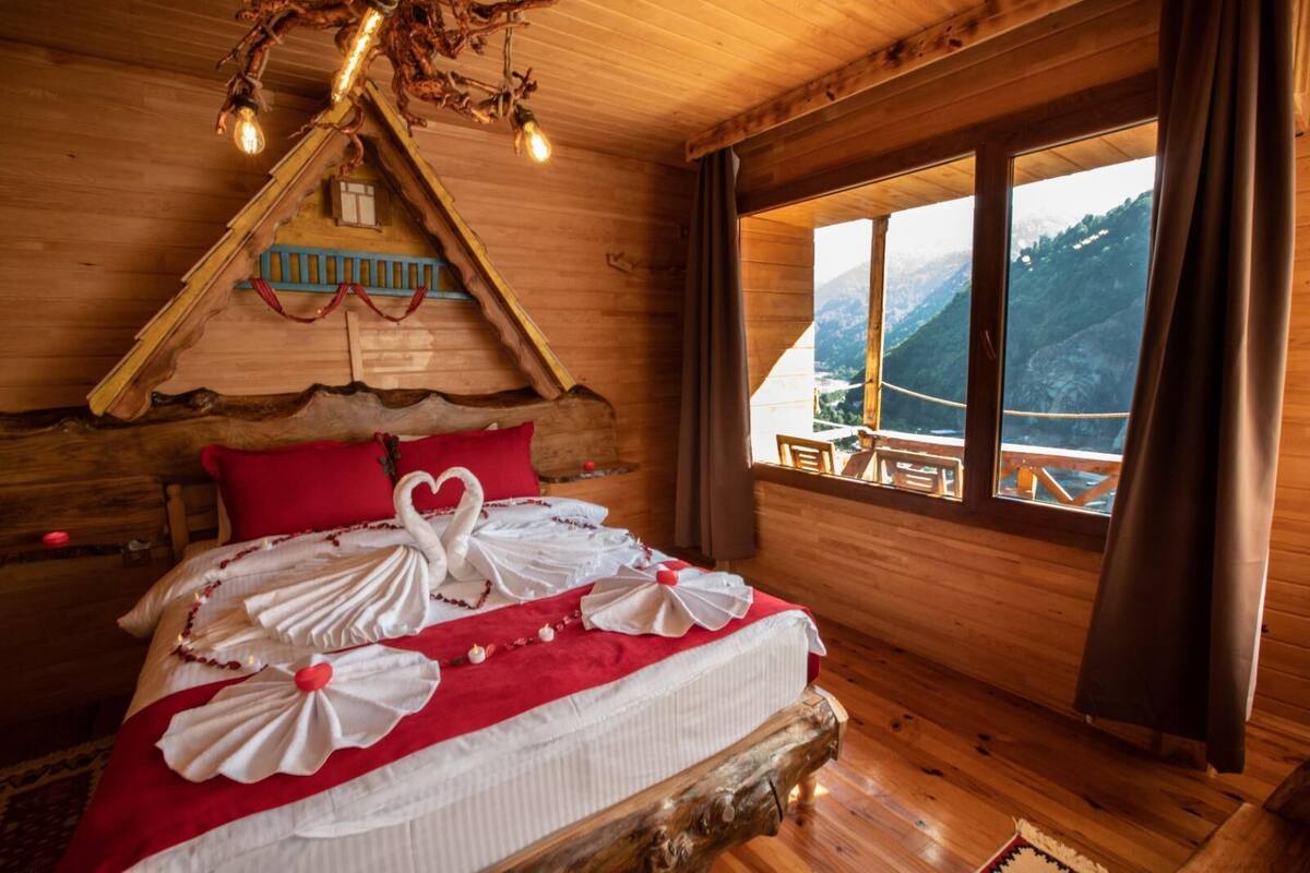 Une chambre confortable dans un chalet en bois, avec un lit décoré de motifs en forme de cœur, un plafond en bois agrémenté de luminaires suspendus et une fenêtre donnant sur une vue imprenable sur les montagnes.