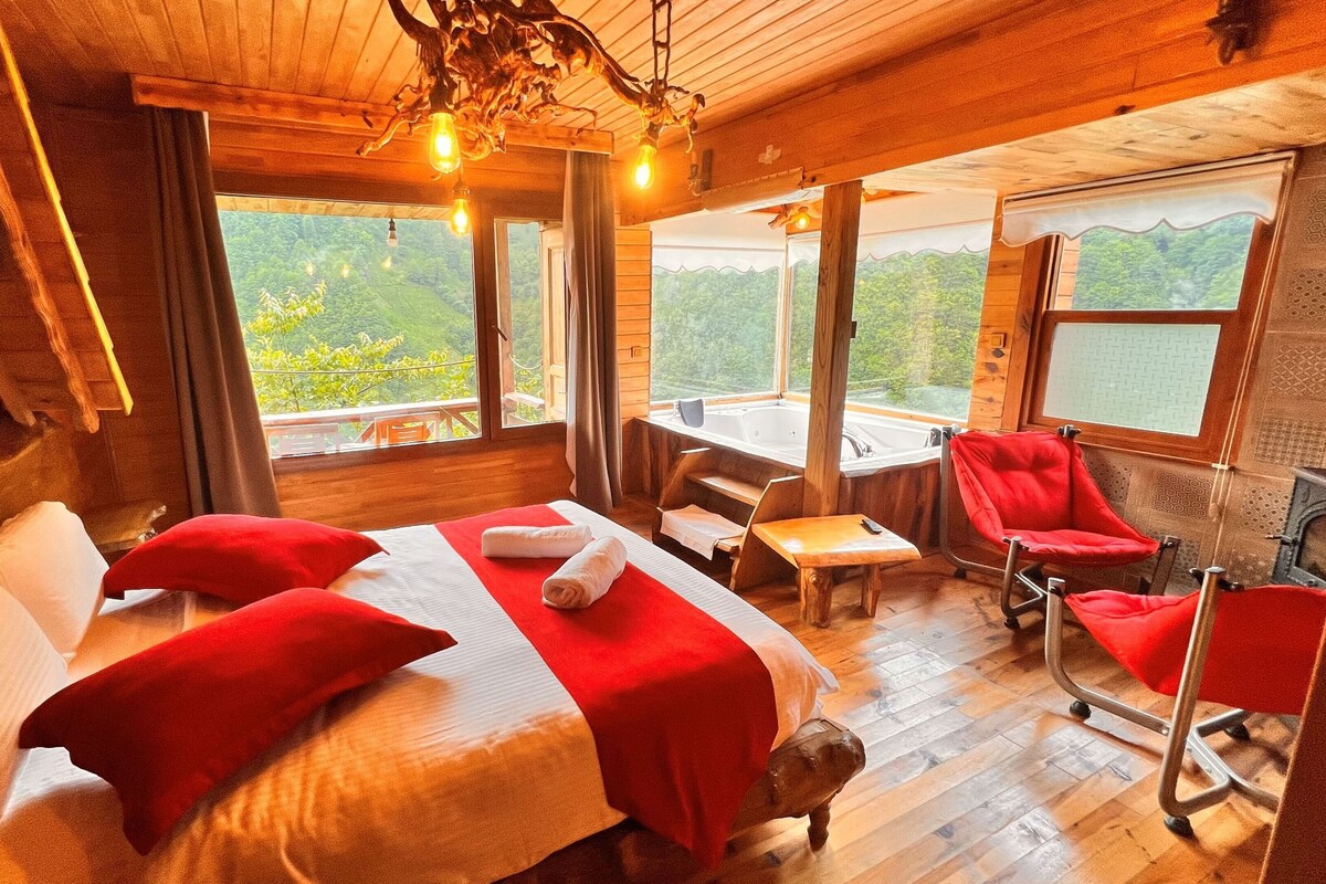 A wooden cabin room with a bed, a desk, a chair, and a view of the outdoors.