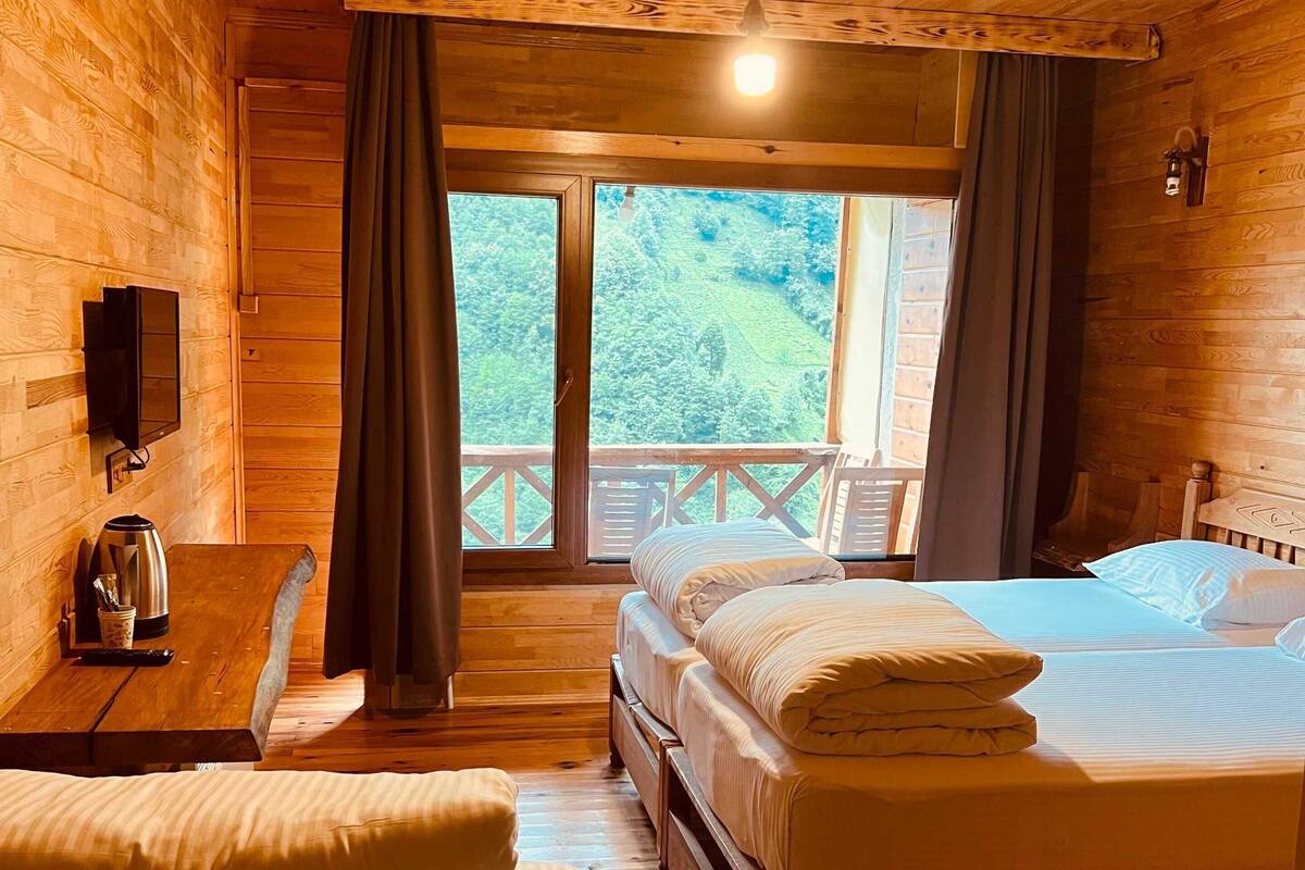 Une chambre confortable de style cabane, avec deux lits, un plafond en bois, une fenêtre avec des rideaux, une petite table avec une bouilloire et un téléviseur fixé au mur.
