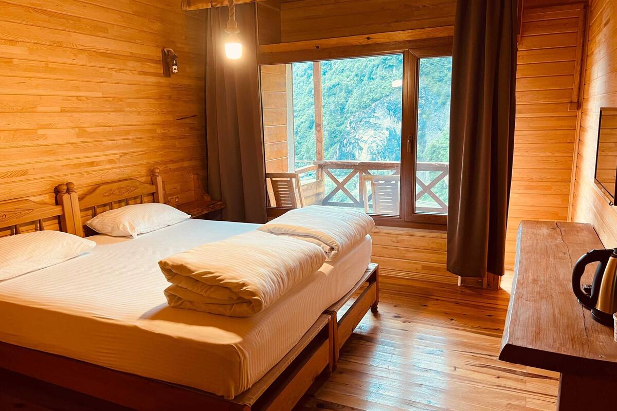 Une chambre dans un chalet en bois, avec deux lits, un bureau en bois et un balcon offrant une vue sur les arbres.