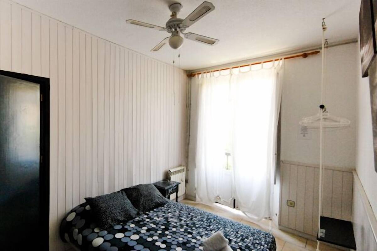 Un dormitorio con cama, ventilador de techo, ventana con cortinas y una puerta.