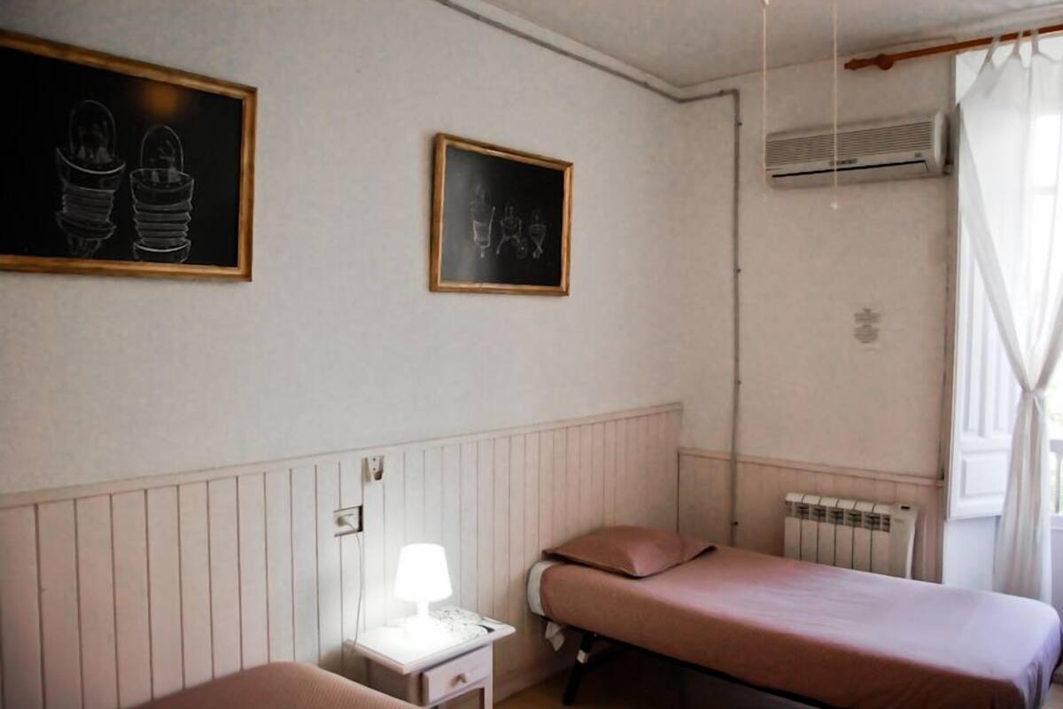 Una habitación con dos camas, una mesita de noche con lámpara, un radiador y dos cuadros en la pared.