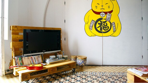 A room with a television on a wooden stand, a bookshelf, a decorative wall with a cat illustration, and a patterned floor. - Itinere Rooms - Hostel (Granada)