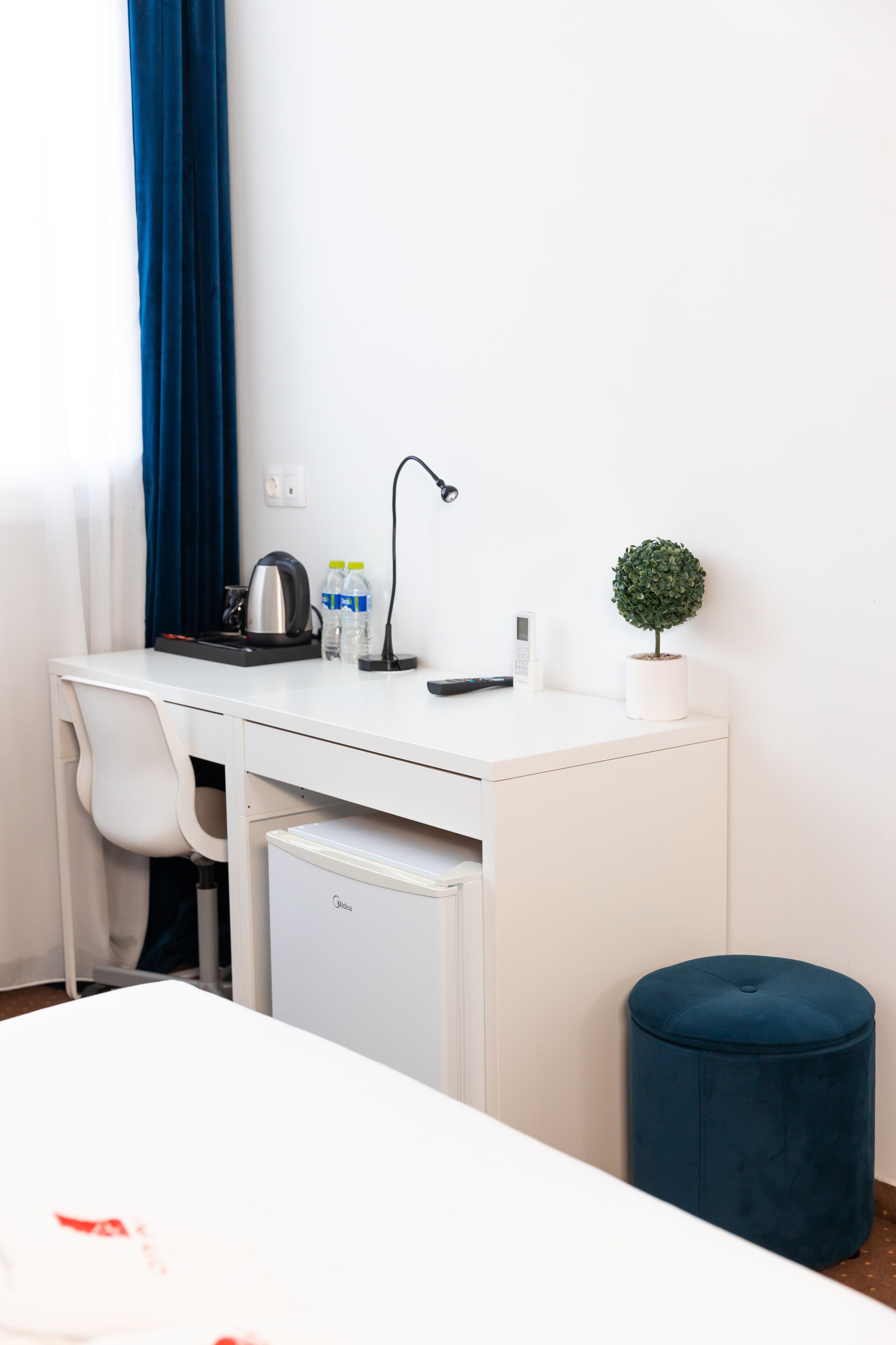 A hotel room with a white desk, a blue ottoman, a small potted plant, a kettle, and a remote control.