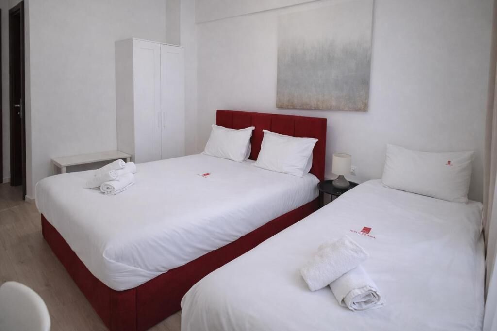 A hotel room with two beds, white bedding, a red headboard, a white wardrobe, and a painting on the wall.