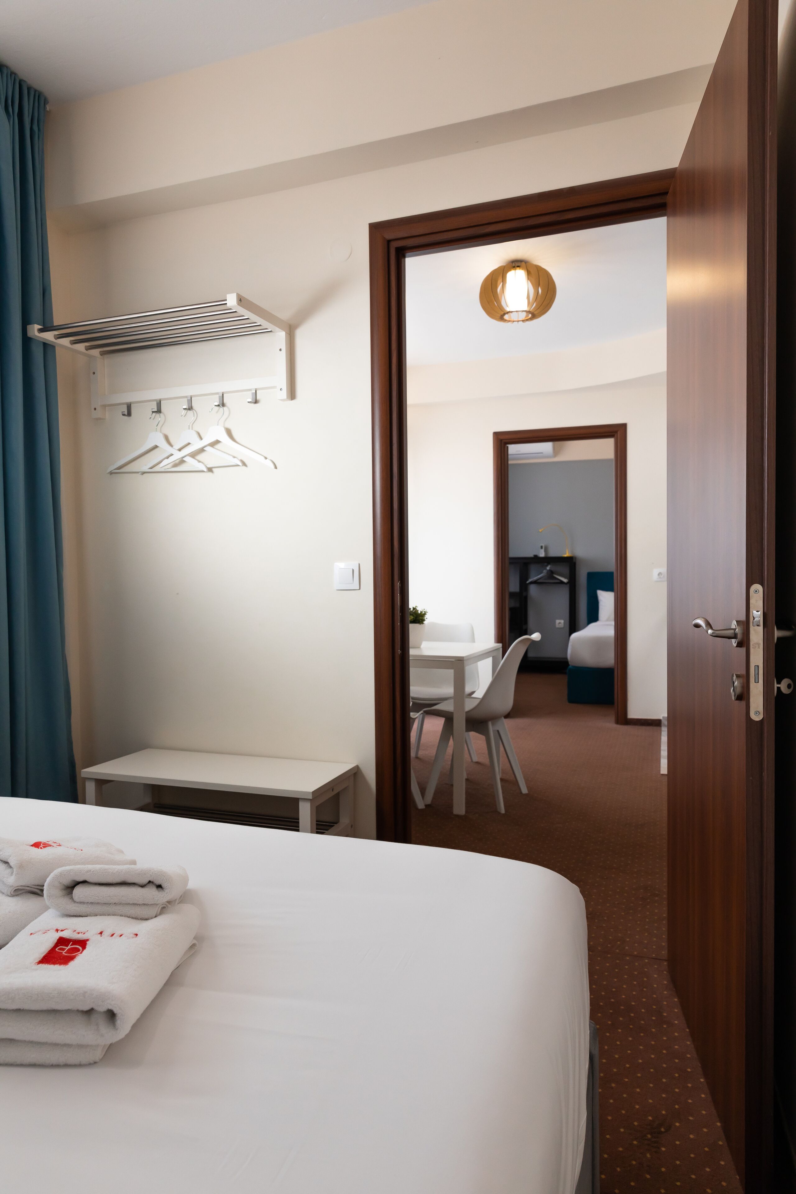 A hotel room with a neatly made bed, a small table with chairs, a hanging clothes rack, and a door leading to a bathroom.