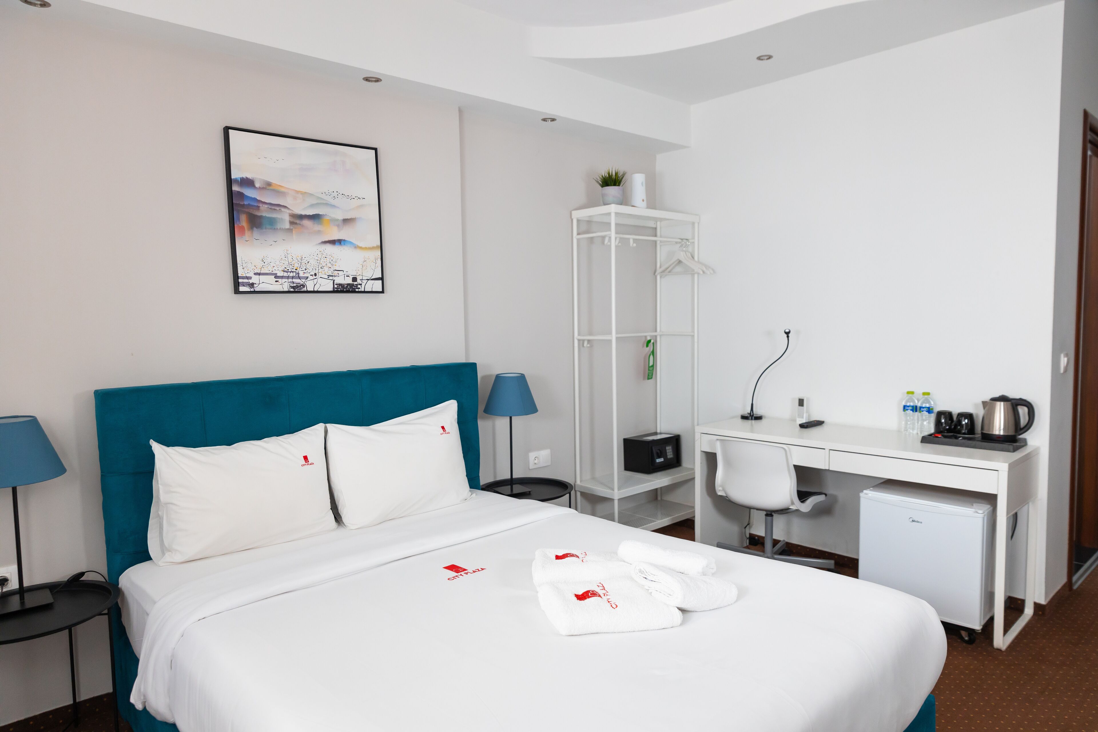 Une chambre d’hôtel avec un lit, un bureau, une chaise, une armoire et un tableau au mur.